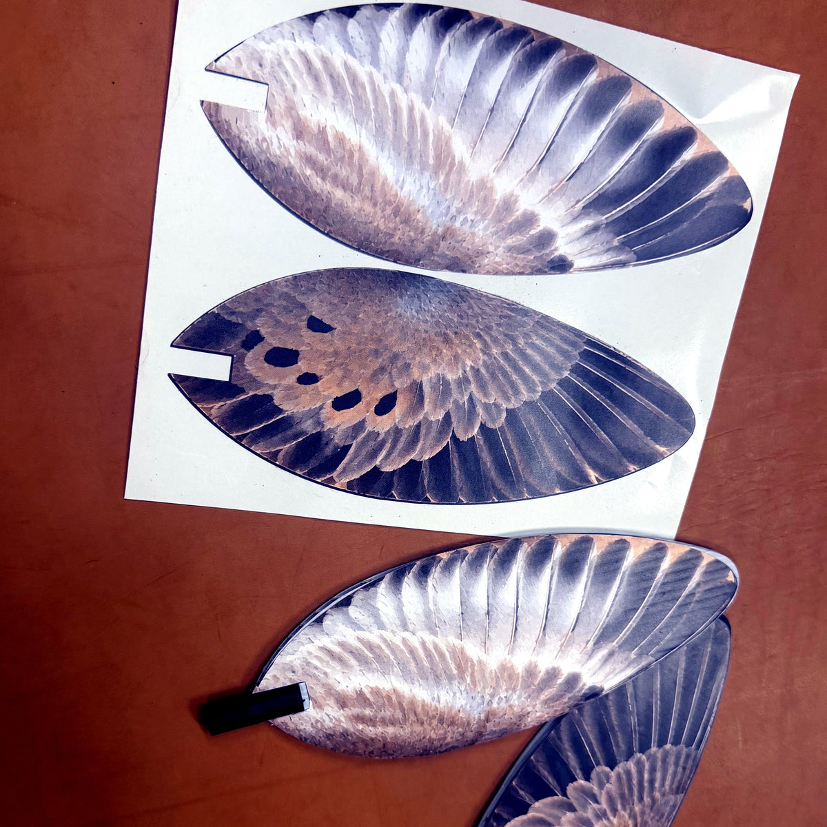 Two Wing Skins vinyl overlay pairs on their printed sheet, close-up on a leather workbench, showing the die-cut edge and mounting notch
